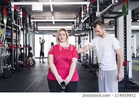 Overweight woman exercising in gym, using kettlebell. Personal trainer couching her and helping her. Overweight woman exercising in gym, using kettlebell. Personal trainer couching her and helping her. 114939952