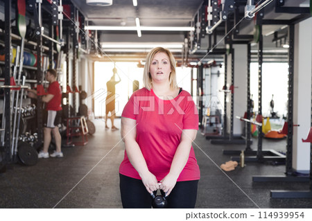 Overweight woman exercising in gym, using kettlebell. Personal trainer couching her and helping her. Overweight woman exercising in gym, using kettlebell. Personal trainer couching her and helping her. 114939954
