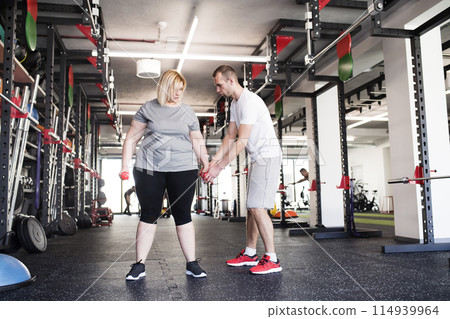 Overweight woman exercising in gym, using dumbbells. Personal trainer couching her and helping her. Overweight woman exercising in gym, using dumbbells. Personal trainer couching her and helping her. 114939964