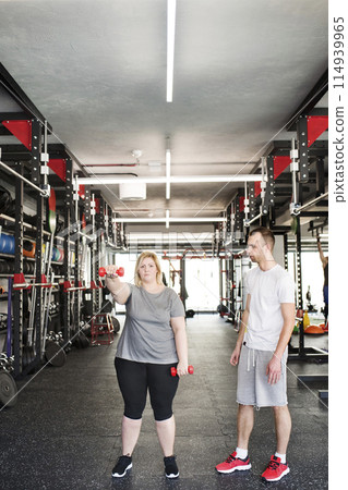 Overweight woman exercising in gym, using dumbbells. Personal trainer couching her and helping her. Overweight woman exercising in gym, using dumbbells. Personal trainer couching her and helping her. 114939965
