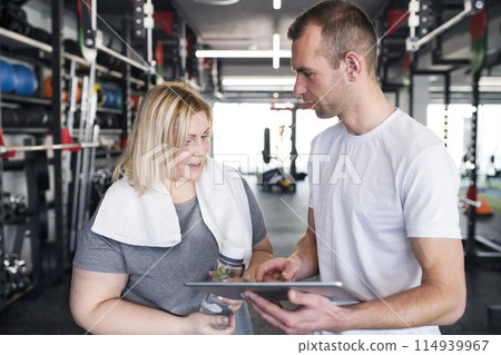 Overweight woman consulting with trainer, discussing daily workout plan. Personal trainer talking with client, discussing exercise program in gym. Overweight woman consulting with trainer, discussing daily workout plan. Personal trainer talking with client, discussing exercise program in gym. 114939967