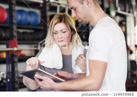 Overweight woman consulting with trainer, discussing daily workout plan. Personal trainer talking with client, discussing exercise program in gym. Overweight woman consulting with trainer, discussing daily workout plan. Personal trainer talking with client, discussing exercise program in gym. 114939968