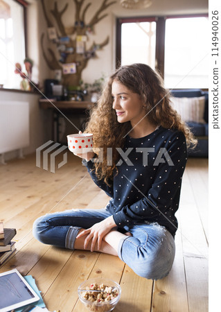 Female student sitting on floor, reading books, studying for final exam at university. Beautiful student at home, drinking coffee. 114940026