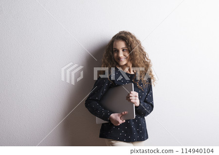 Portrait of a gorgeous teenage girl with curly hair, holding notebook, leaning against wall. Studio shot, white background with copy space Portrait of a gorgeous teenage girl with curly hair, holding notebook, leaning against wall. Studio shot, white background with copy space 114940104
