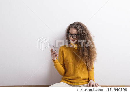 Portrait of a gorgeous teenage girl with curly hair, holding smartphone, scrolling. Studio shot, white background with copy space 114940108