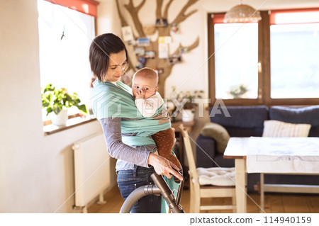 Mother holding small baby while vacuuming floor, carrying him around house in baby wrap. Household chores. Mother holding small baby while vacuuming floor, carrying him around house in baby wrap. Household chores. 114940159