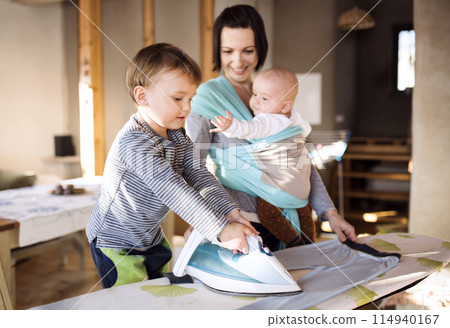 Mother carrying small baby in wrap, older son helping with ironing. Family doing household chores. Mother carrying small baby in wrap, older son helping with ironing. Family doing household chores. 114940167