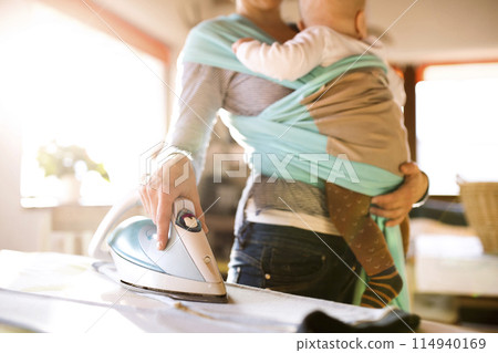 Mother holding small baby while ironing, carrying him around house in baby wrap. Unconditional paternal love, mother's Day concept. Mother holding small baby while ironing, carrying him around house in baby wrap. Unconditional paternal love, mother's Day concept. 114940169
