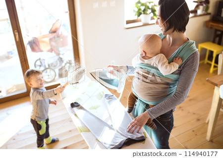 Mother carrying small baby in wrap, older son helping with celaning home, ironing. Family doing household chores. Mother carrying small baby in wrap, older son helping with celaning home, ironing. Family doing household chores. 114940177