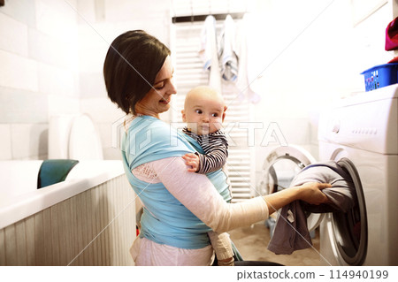 Mother holding small baby while loading the washing machine with dirty laundry, carrying him around house in baby wrap. Household chores. Mother holding small baby while loading the washing machine with dirty laundry, carrying him around house in baby wrap. Household chores. 114940199