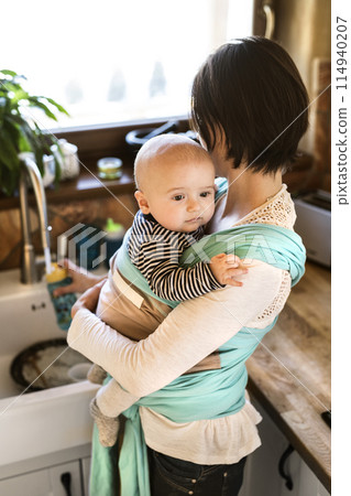 Mother holding small baby while cleaning dishes, carrying him around house in baby wrap. Household chores. Mother holding small baby while cleaning dishes, carrying him around house in baby wrap. Household chores. 114940207
