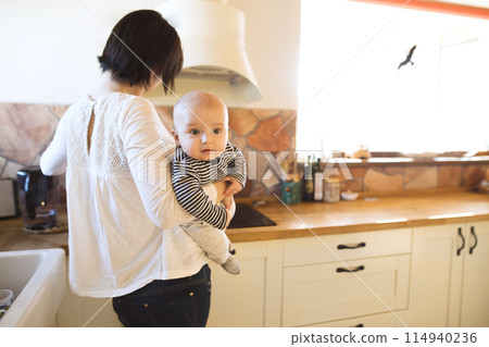 Mother holding small baby, carrying him around house while cooking. Unconditional paternal love, mother's Day concept. Mother holding small baby, carrying him around house while cooking. Unconditional paternal love, mother's Day concept. 114940236