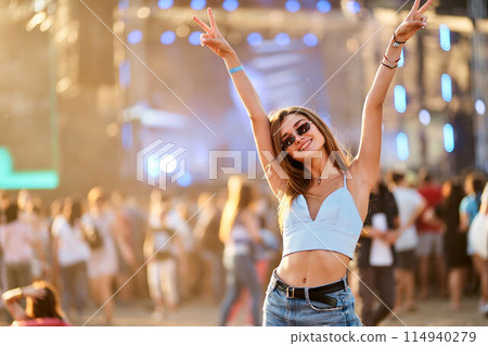 Happy young woman at outdoor music festival raises peace signs, enjoys summer event on sunny day. Crowd dances, socializes at beach concert. Fashionable festivalgoer lives up moment, carefree vibes. Happy young woman at outdoor music festival raises peace signs, enjoys summer event on sunny day. Crowd dances, socializes at beach concert. Fashionable festivalgoer lives up moment, carefree vibes. 114940279