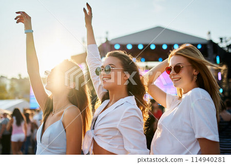 Group of joyful young women dance at sunny beach music festival. Girls in summer outfits enjoy concert, hands up in celebration. Vibrant fest atmosphere, friends share fun experience by sea. 114940281