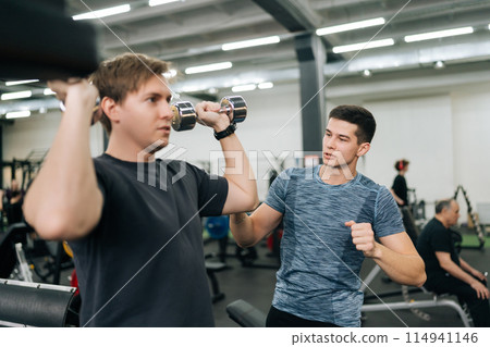 Closeup of beginner sportsman working out with dumbbell with personal fitness trainer at gym. Sporty guy doing weight exercises with assistance of coach at sport club. Concept of healthy lifestyle. 114941146