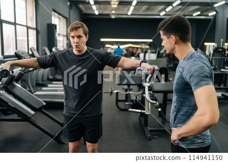 Handsome beginner sportsman doing side lateral raise with dumbbells with personal fitness trainer at gym. Sporty guy doing weight exercises with assistance of coach. Concept of healthy lifestyle. 114941150