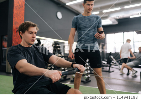 Focused beginner sportsman with personal instructor working out in gym by using rowing machine. Sweating man doing exercises for back muscles under supervision of trainer measuring time on stopwatch. Focused beginner sportsman with personal instructor working out in gym by using rowing machine. Sweating man doing exercises for back muscles under supervision of trainer measuring time on stopwatch. 114941188