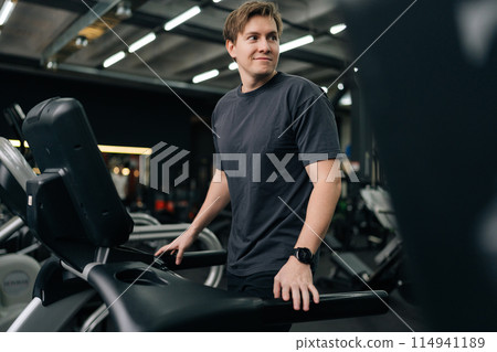 Smiling young man in sportswear walking on treadmill preforming warming-up run routine exercise in gym. Sporty male walking on running track, working on health and fitness. Concept of healthy life. Smiling young man in sportswear walking on treadmill preforming warming-up run routine exercise in gym. Sporty male walking on running track, working on health and fitness. Concept of healthy life. 114941189
