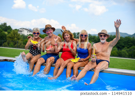 Group of cheerful seniors sitting on edge of pool with legs in water, eating watermelon. Elderly friends spending hot day by swimming pool, sunbathing. Group of cheerful seniors sitting on edge of pool with legs in water, eating watermelon. Elderly friends spending hot day by swimming pool, sunbathing. 114941510