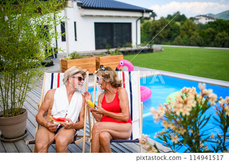 Beautiful elderly couple enjoying their vacation. Seniors having romantic time, sitting by swimming pool in lounche chairs eating watermelon. 114941517