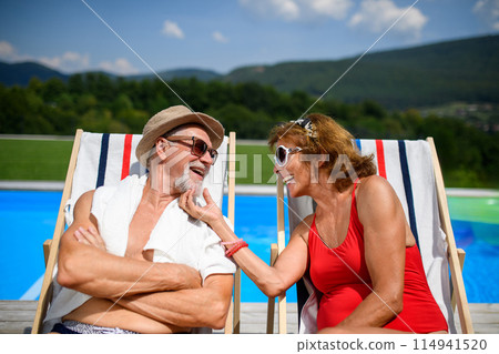 Beautiful elderly couple enjoying their vacation. Seniors having romantic time, sitting by swimming pool in lounche chairs sunbathing. 114941520