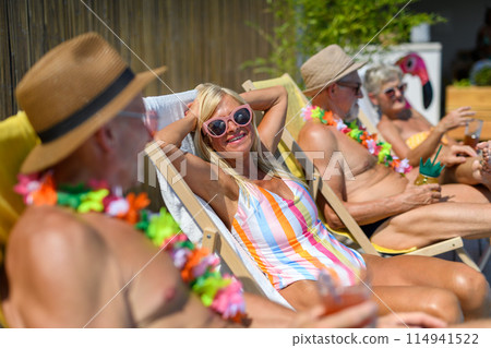 Group of cheerful seniors sitting lounge chairs by a swimming pool and sunbathing. Beautiful senior woman enjoying a hot day outdoors in backyard with friends. 114941522