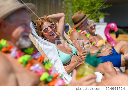 Group of cheerful seniors sitting lounge chairs by a swimming pool and sunbathing. Beautiful senior woman enjoying a hot day outdoors in backyard with friends. 114941524