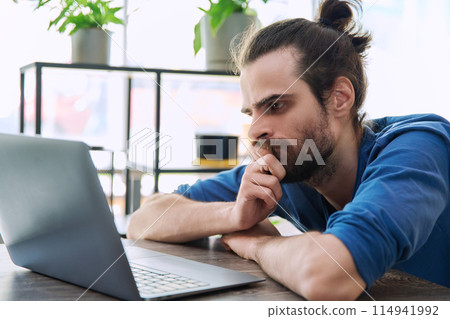 Serious tired young man looking at laptop screen Serious tired young man looking at laptop screen 114941992