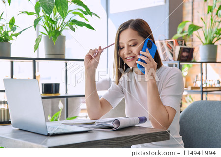 Young woman sitting at table with laptop talking on mobile phone in coworking Young woman sitting at table with laptop talking on mobile phone in coworking 114941994