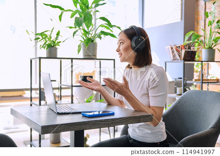 Young woman in headphones having video chat conference using laptop sitting in cafe Young woman in headphones having video chat conference using laptop sitting in cafe 114941997