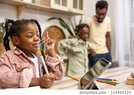 African American child siting at table completing her schoolwork with the help of her digital tablet 114942124
