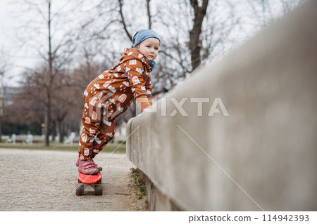 Cute toddler girl playing outdoor in playground with skateboard. Girl in softshell bodysuit spending time in park. 114942393