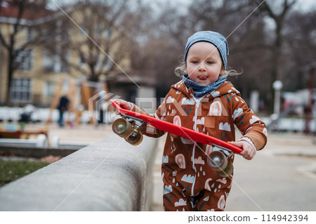 Cute toddler girl playing outdoor in playground with skateboard. Girl in softshell bodysuit spending time in park. 114942394