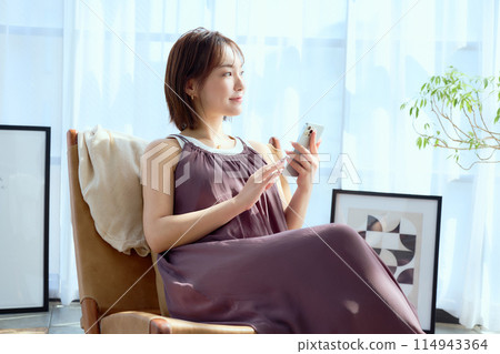 A woman sitting on a chair in a room and looking at her smartphone 114943364