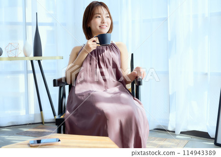 A woman sitting on a chair in a room and drinking coffee 114943389