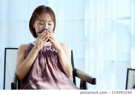 A woman sitting on a chair in a room and drinking coffee 114943398