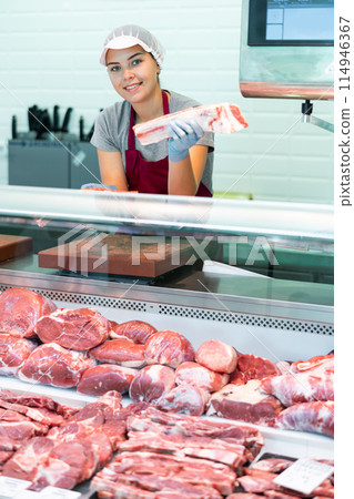 Young female seller shows demonstrates raw beef ribs in glass showcase of refrigerator. Young female seller shows demonstrates raw beef ribs in glass showcase of refrigerator. 114946367
