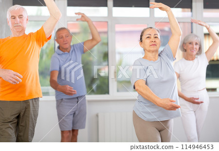 Happy aged woman in sportswear confidently leading ballet class with dancers following movements in dance studio Happy aged woman in sportswear confidently leading ballet class with dancers following movements in dance studio 114946453