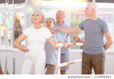 Group of elderly dancers engaged in ballet in dance studio perfoms exercise near ballet barre, senior male choreographer helps to coordinate movements correctly 114946461