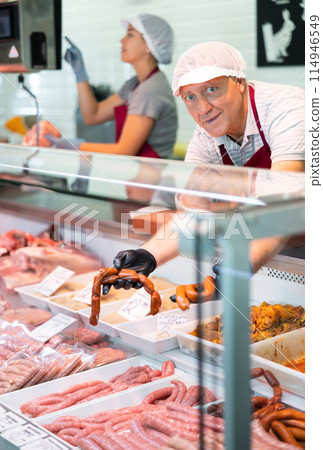 Adult man seller lays out sausages in butcher shop 114946549