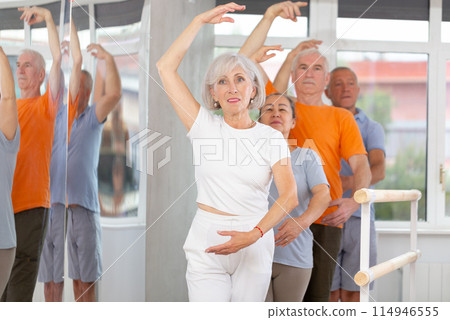 Ordinary elderly people stand in third position near ballet barre during group training in dance studio Ordinary elderly people stand in third position near ballet barre during group training in dance studio 114946555