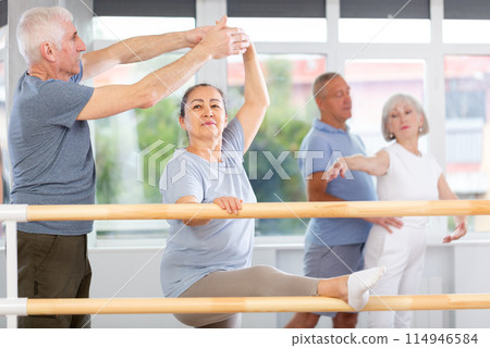Active mature woman practicing leg stretching holding by ballet barre with instructor 114946584