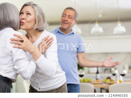 Two elderly woman greet each other by kissing each other on the cheek. While men shake hands Two elderly woman greet each other by kissing each other on the cheek. While men shake hands 114946585