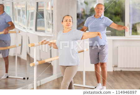Active mature woman practicing battement tendu holding by ballet barre together with others 114946897