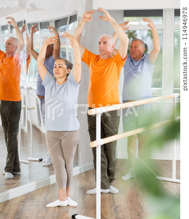 Active mature woman practicing fifth position holding by ballet barre together with others 114946978