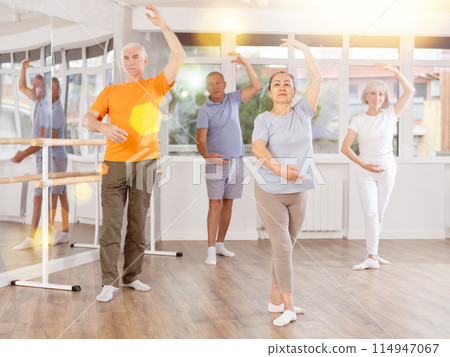 Happy aged woman in sportswear confidently leading ballet class with dancers following movements in dance studio 114947067