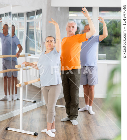 Ordinary elderly people stand in third position near ballet barre during group training in dance studio 114947069