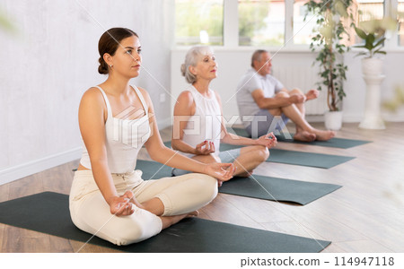Women sitting on mats in Lotus pose Padmasana during yoga stretch joint workout in fitness center 114947118