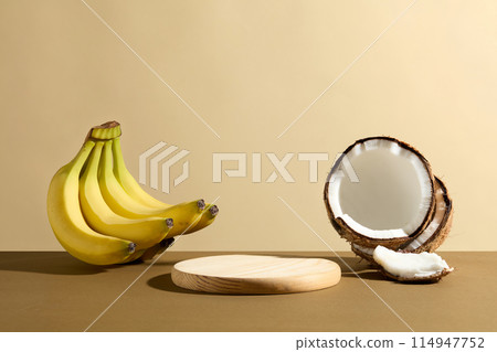 Advertising photo with a wooden pedestal in round shaped displayed on brown table next to some coconut and a bunch of banana, over light yellow background. Vacant space for design or adding text Advertising photo with a wooden pedestal in round shaped displayed on brown table next to some coconut and a bunch of banana, over light yellow background. Vacant space for design or adding text 114947752