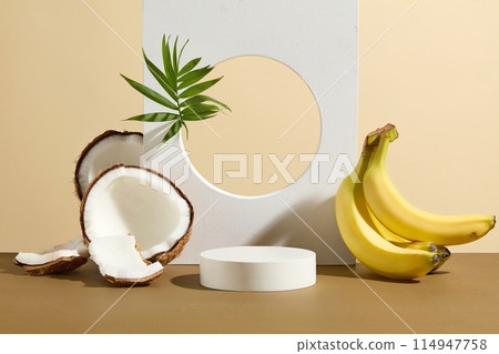 The white backdrop has a hole in center striking with a green tree branch, white geometric podium next to a bunch of yellow banana and some pieces of coconut featured on light yellow background 114947758